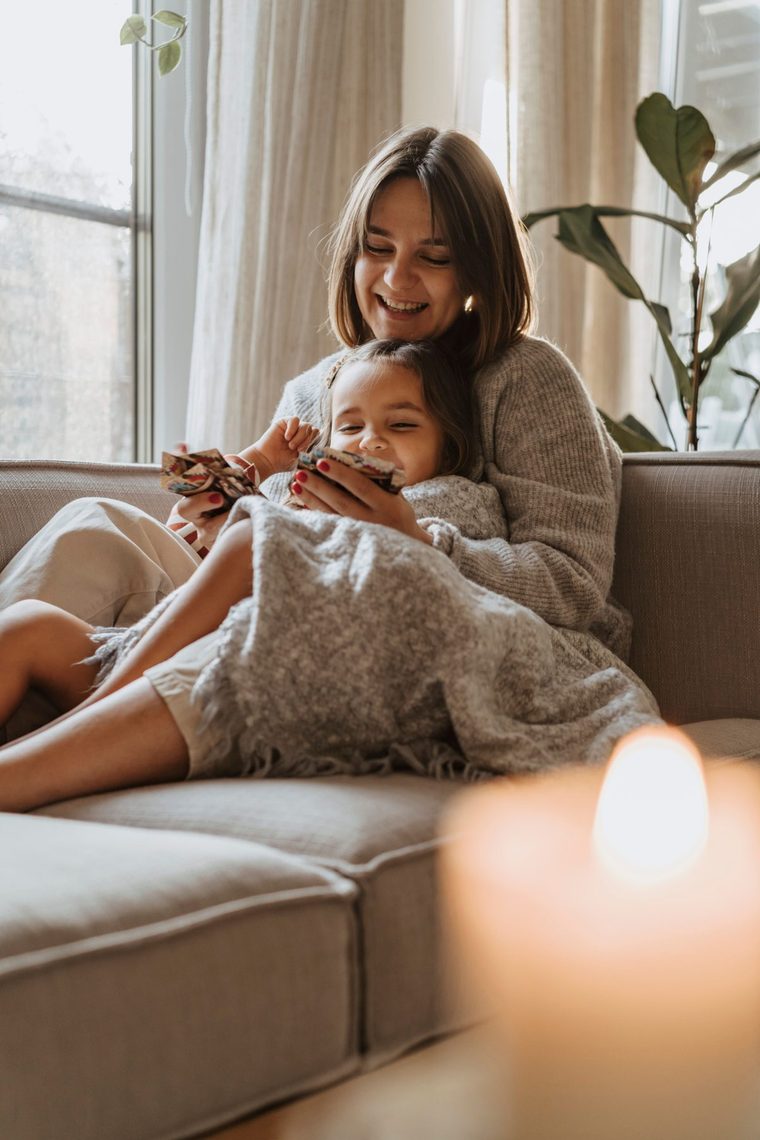 Uśmiechnięta mama z córką pod kocem na kanapie w jasnym salonie, oglądają razem kolorową książeczkę, z przodu pali się świeca